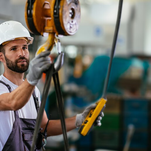 A crane operator wearing a hard hat holding a crane hook and a pendant controller in a manufacturing factory.