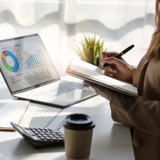 Woman writing in a notebook and sitting in front of a laptop with an analytics dashboard.